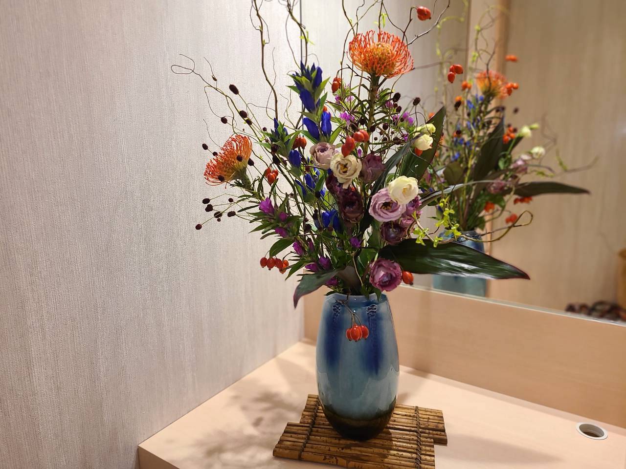 洗面スペースの傍らに飾られた生け花。青い花器に、のびやかな枝と季節の花や実ものが大ぶりに生けられている。