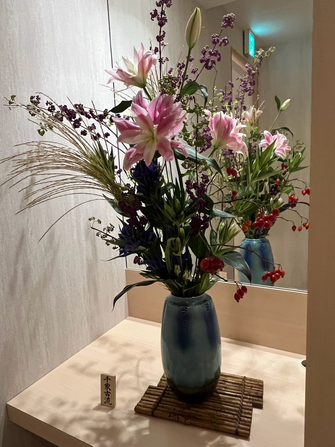 店内に飾られた新しい生け花。ピンクの花々と枝物を青緑の花器に生けた様子
